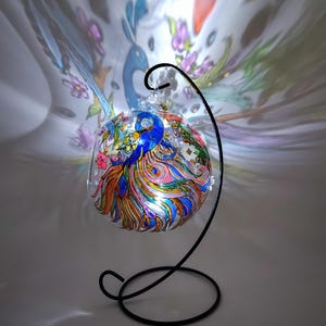 K&ouml;nnte beinhalten: Ein gl&auml;serner Weihnachtsbaumschmuck mit einem Pfauendesign, das in Blau, Rosa, Gelb und Gr&uuml;n bemalt ist. Der Schmuck wird von innen beleuchtet und steht auf einem schwarzen Metallst&auml;nder.