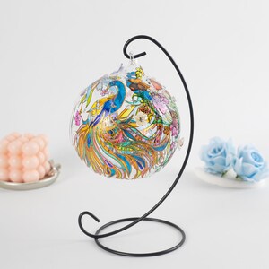 K&ouml;nnte beinhalten: Ein Glasornament mit einem Pfauendesign, das in leuchtenden Farben wie Blau, Gr&uuml;n, Gelb und Pink bemalt ist. Das Ornament steht auf einem schwarzen Metallst&auml;nder.