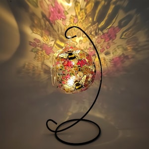 May include: A glass globe with a floral and bee design hangs from a black metal stand. The globe is illuminated from within, casting a warm glow.