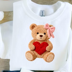 Puede incluir: Sudadera blanca con diseño de oso de peluche. El oso de peluche marrón tiene un lazo rosa y sostiene un corazón rojo. La sudadera tiene cuello redondo y mangas largas.