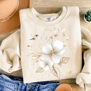 Puede incluir: Sudadera color crema con un diseño floral de flores blancas, hojas marrones y un colibrí. El diseño incluye una pequeña mariposa y detalles de gotas de agua. La sudadera se combina con vaqueros azules y sandalias marrones.
