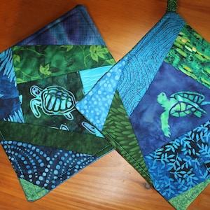 May include: Two blue and green quilted pot holders with a sea turtle design. The pot holders are made of fabric scraps and have a patchwork design.