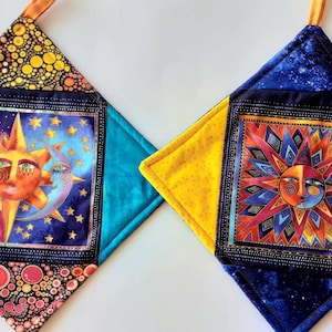 May include: Two quilted pot holders with a diamond shape. One pot holder has a blue, yellow, and black background with a sun and moon design. The other pot holder has a yellow, blue, and black background with a sun design.