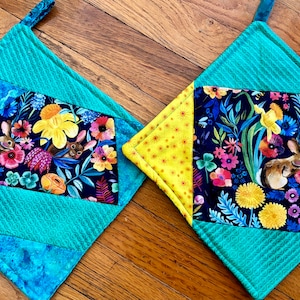May include: Two quilted pot holders with a floral pattern featuring bunnies and yellow flowers. The pot holders are made of a patchwork of different fabrics in shades of blue, green, yellow, and black.