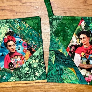 May include: Two green quilted pot holders with a diamond-shaped design. Each pot holder features a portrait of Frida Kahlo in the center. One pot holder has a green and white floral pattern, while the other has a green and white leaf pattern.