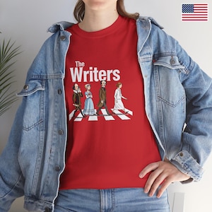 Puede incluir: Camiseta roja con la frase "The Writers" y una ilustraci&oacute;n de escritores famosos cruzando un paso de cebra. Los escritores incluyen a William Shakespeare y Jane Austen. La camiseta se lleva con una chaqueta vaquera azul.