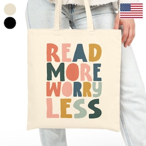 Könnte beinhalten: Eine cremefarbene Tragetasche mit dem Schriftzug "READ MORE WORRY LESS" in bunten, fetten Buchstaben. Die Tasche hat zwei Henkel und wird von einer Person gehalten. Der Text ist in Orange-, Blau-, Rosa-, Gelb- und Grüntönen gehalten.