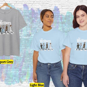 Puede incluir: Camisetas gris deportivo y azul claro con el texto "The Writers" y un gr&aacute;fico de personas cruzando una calle. Dos personas llevan camisetas azul claro. Una camiseta gris deportivo cuelga de una percha de madera.