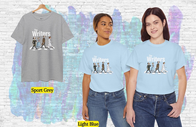 Puede incluir: Camisetas gris deportivo y azul claro con el texto "The Writers" y un gr&aacute;fico de personas cruzando una calle. Dos personas llevan camisetas azul claro. Una camiseta gris deportivo cuelga de una percha de madera.