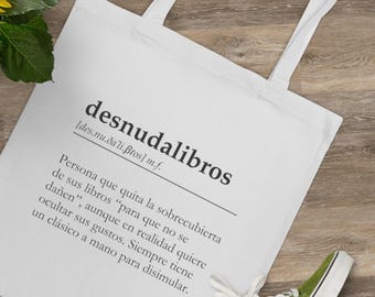 May include: White tote bag with a black graphic design that defines the word "desnudalibros" as a person who removes the dust jacket from their books to avoid damaging them, but in reality, they are hiding their true tastes. The bag is lying on a wooden surface.