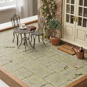 Puede incluir: Una escena de patio en miniatura con una mesa redonda de metal y dos sillas a juego, un pequeño olivo en maceta, una planta de rosas trepadoras contra una pared de ladrillo, un felpudo y una regadera color cobre junto a una puerta doble.