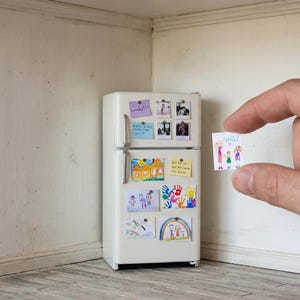 Może przedstawiać: Miniaturowa biała lodówka ze zdjęciami i rysunkami przymocowanymi magnesami. Ręka trzyma mały rysunek ze słowem "Rodzina" i postaciami z patyków. Lodówka znajduje się w małym pokoju z białymi ścianami.