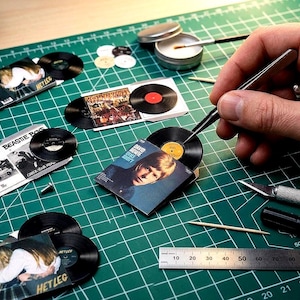 May include: Miniature vinyl record replicas and album covers featuring David Bowie's Blackstar, Beastie Boys, and Wet Leg. The collection includes small black discs and square paper sleeves with printed artwork, displayed on a green cutting mat.