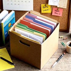May include: A brown corrugated cardboard storage box containing multiple hanging file folders in shades of purple, red, green, and yellow. The box features a cutout handle on the front and is filled with documents, sitting on a wooden desk surface.