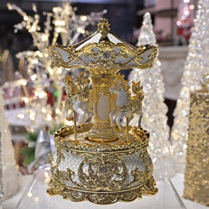 May include: A gold and white carousel music box with three white horses. The carousel has ornate detailing and is surrounded by Christmas decorations, including white Christmas trees and a gold sequined gift box.