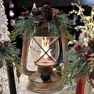 Puede incluir: Una linterna de metal r&uacute;stico con un recinto de vidrio transparente, decorada con pi&ntilde;as, bayas rojas y ramitas de hoja perenne. La linterna tiene un brillo c&aacute;lido y acogedor, perfecto para la decoraci&oacute;n navide&ntilde;a.