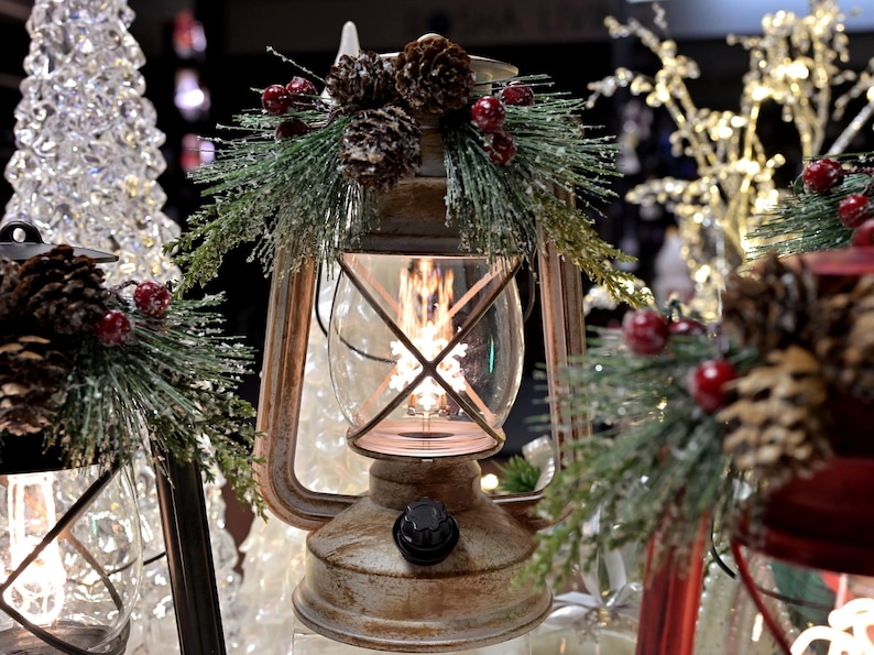 Puede incluir: Una linterna de metal r&uacute;stico con un recinto de vidrio transparente, decorada con pi&ntilde;as, bayas rojas y ramitas de hoja perenne. La linterna tiene un brillo c&aacute;lido y acogedor, perfecto para la decoraci&oacute;n navide&ntilde;a.