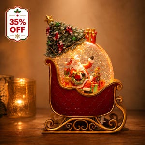 May include: A decorative Christmas snow globe shaped like a red sleigh with gold scrollwork runners. Inside the globe, a Santa Claus figure sits surrounded by gifts. A decorated Christmas tree with a gold star sits atop the sleigh.