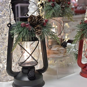 Puede incluir: Linterna de metal negro con una carcasa de vidrio, decorada con pi&ntilde;as, bayas artificiales y vegetaci&oacute;n. Tambi&eacute;n se ve una linterna roja con una silueta de Pap&aacute; Noel. Decoraci&oacute;n navide&ntilde;a.