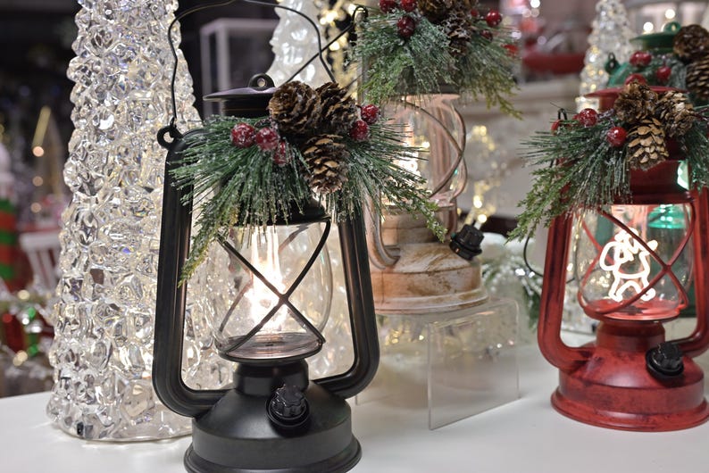 Puede incluir: Linterna de metal negro con una carcasa de vidrio, decorada con pi&ntilde;as, bayas artificiales y vegetaci&oacute;n. Tambi&eacute;n se ve una linterna roja con una silueta de Pap&aacute; Noel. Decoraci&oacute;n navide&ntilde;a.