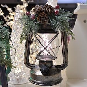 Puede incluir: Una linterna decorativa con un globo de cristal transparente y un marco de metal. La linterna est&aacute; adornada con agujas de pino, pi&ntilde;as y bayas rojas. La linterna tiene un asa y un bot&oacute;n en la base. El tema general es la decoraci&oacute;n navide&ntilde;a.