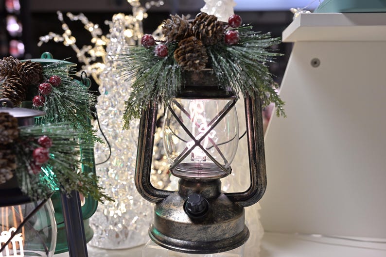 Puede incluir: Una linterna decorativa con un globo de cristal transparente y un marco de metal. La linterna est&aacute; adornada con agujas de pino, pi&ntilde;as y bayas rojas. La linterna tiene un asa y un bot&oacute;n en la base. El tema general es la decoraci&oacute;n navide&ntilde;a.