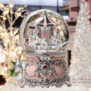 May include: A pink and silver carousel snow globe with a clear glass dome. The carousel features white horses and a pink and silver canopy. The base is pink with ornate silver detailing and a red jewel. Text on the base reads "6.8-7.5" Pink & Silver Carousel Collection."