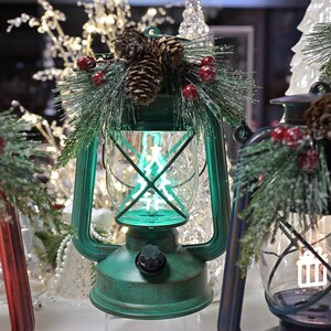 Puede incluir: Faroles decorativos rojos, verdes y azules, cada uno adornado con pi&ntilde;as, ramas de pino artificiales y bayas rojas. Los faroles tienen un estilo vintage y est&aacute;n iluminados con un suave brillo verde.