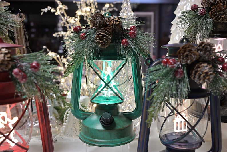 Puede incluir: Faroles decorativos rojos, verdes y azules, cada uno adornado con pi&ntilde;as, ramas de pino artificiales y bayas rojas. Los faroles tienen un estilo vintage y est&aacute;n iluminados con un suave brillo verde.