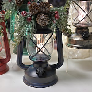Puede incluir: Una colecci&oacute;n de faroles decorativos en varios colores, incluyendo rojo, azul y bronce. Cada farol tiene una carcasa de vidrio transparente con una luz decorativa en el interior. Algunos est&aacute;n adornados con pi&ntilde;as y vegetaci&oacute;n, sugiriendo un tema festivo.