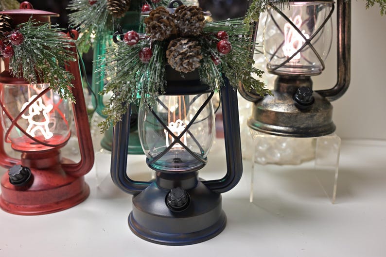 Puede incluir: Una colecci&oacute;n de faroles decorativos en varios colores, incluyendo rojo, azul y bronce. Cada farol tiene una carcasa de vidrio transparente con una luz decorativa en el interior. Algunos est&aacute;n adornados con pi&ntilde;as y vegetaci&oacute;n, sugiriendo un tema festivo.