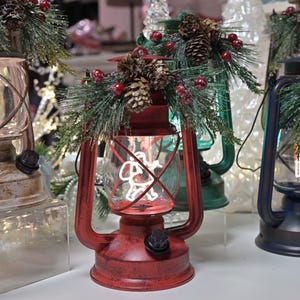 Puede incluir: Varias linternas decorativas rojas, verdes y azules. Cada linterna tiene una carcasa de vidrio transparente con un dise&ntilde;o festivo y est&aacute; adornada con pi&ntilde;as, vegetaci&oacute;n y bayas rojas. Las linternas est&aacute;n iluminadas, creando un brillo c&aacute;lido y acogedor.
