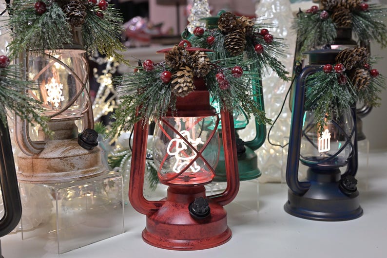 Puede incluir: Varias linternas decorativas rojas, verdes y azules. Cada linterna tiene una carcasa de vidrio transparente con un dise&ntilde;o festivo y est&aacute; adornada con pi&ntilde;as, vegetaci&oacute;n y bayas rojas. Las linternas est&aacute;n iluminadas, creando un brillo c&aacute;lido y acogedor.