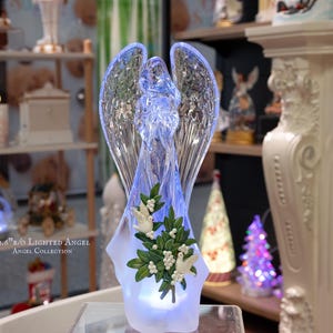 May include: A 12.6-inch lighted angel figurine with clear wings and a white base. The angel holds a bouquet of white flowers and green leaves. The text "12.6" H/O Lighted Angel Angel Collection" is visible on the base.