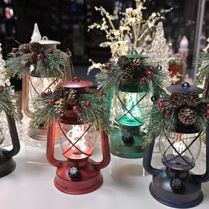 Puede incluir: Una colecci&oacute;n de faroles navide&ntilde;os decorativos en varios colores, incluyendo rojo, verde y negro. Cada farol est&aacute; adornado con pi&ntilde;as, vegetaci&oacute;n y bayas rojas. Los faroles tienen un dise&ntilde;o vintage con un recinto de vidrio transparente y un brillo c&aacute;lido.