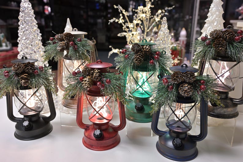Puede incluir: Una colecci&oacute;n de faroles navide&ntilde;os decorativos en varios colores, incluyendo rojo, verde y negro. Cada farol est&aacute; adornado con pi&ntilde;as, vegetaci&oacute;n y bayas rojas. Los faroles tienen un dise&ntilde;o vintage con un recinto de vidrio transparente y un brillo c&aacute;lido.