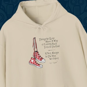 May include: A beige hoodie with a red and white checkered pattern on the front. The hoodie has a quote that reads "Things We Lose Have A Way Of Coming Back To Us In The End If Not Always In The Way We Expect".