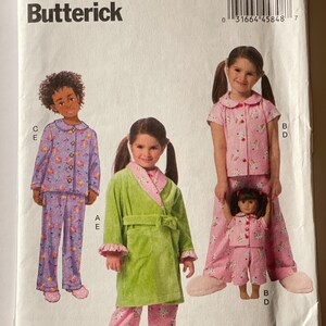 May include: A sewing pattern for children's pajamas and a robe. The pattern includes a girl wearing a pink and white pajama set with a unicorn print, a girl wearing a purple and white pajama set with a rainbow print, and a girl wearing a green robe with a pink and white pajama set with a unicorn print. The pattern also includes a doll wearing a pink and white pajama set with a unicorn print. The pattern number is B6123.