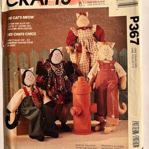May include: A McCall's Crafts sewing pattern envelope labeled P367 featuring illustrations of three handmade cat dolls wearing fabric clothing. The text includes The Cat's Meow, Tom and Allie Cat, and by Faye Wine. The dolls are 21 inches tall.