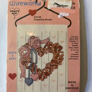 Pode incluir: Um kit de ponto cruz com uma coroa de videira com um coração no centro. O kit inclui um cabide de arame preto, tecido Aida de 14 pontos, uma agulha, fio e um coração para cortar. A embalagem é rosa e branca com as palavras "Country Wireworks" e "What's New, Inc. Mesa, AZ USA".