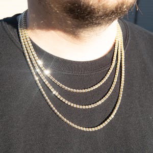 May include: A layered gold chain necklace with small, clear stones. The necklace is worn by a person with a black shirt.