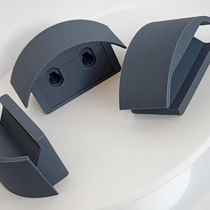 May include: Three dark gray, curved, semi-circular mounting brackets with smooth surfaces. Each bracket features a flat base with two circular keyhole slots for wall attachment and a wide, arched top section designed to hold or support objects.