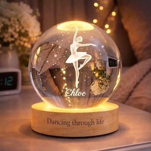 Puede incluir: Una esfera de cristal iluminada con una bailarina grabada en su interior, sobre una base de madera. La base dice "Dancing through life" y el nombre "Chloe" está grabado en la esfera. La luz cálida crea un ambiente acogedor.