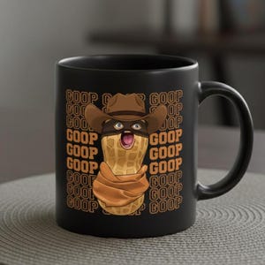 May include: Black ceramic mug featuring a cartoon peanut character wearing a cowboy hat and mask. The peanut is wrapped in a brown scarf. The word "GOOP" is repeated in a pattern around the character. The mug is on a gray placemat.