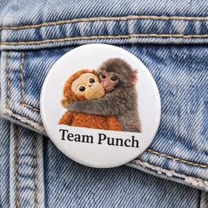 May include: A white button pin with the text "Team Punch" features a brown stuffed monkey hugging a gray monkey. The pin is attached to a blue denim jacket.