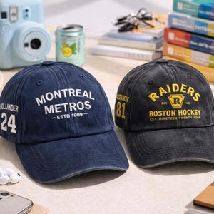 Könnte beinhalten: Zwei Baseballkappen. Eine ist marineblau mit "MONTREAL METROS - ESTD 1909" in Weiß und "HOLLANDER 24" an der Seite. Die andere ist schwarz mit "RAIDERS BOSTON HOCKEY EST. NINETEEN TWENTY-FOUR" in Gelb und "ROZANOV 81" an der Seite.