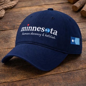 May include: A navy blue baseball cap featuring the embroidered word "minnesota" in white, with the phrases "human decency & hotdish." A small flag patch is on the side. The cap is made of a soft fabric.
