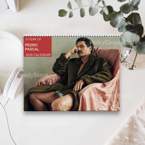 May include: A spiral-bound calendar featuring a photo of Pedro Pascal. The calendar has a red banner with the text "A YEAR OF PEDRO PASCAL 2026 CALENDAR". The image shows Pascal in a green coat, seated in a pink chair.