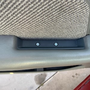 May include: Close-up of a car door interior. The image shows a section of the door panel with a gray fabric section and a light gray plastic trim. A black plastic piece is attached with two silver screws.
