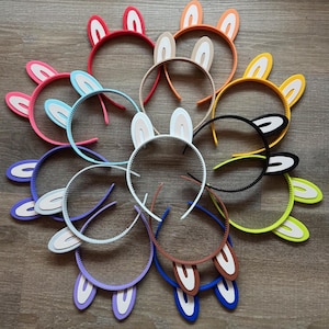 May include: A collection of colorful bunny ear headbands. The headbands feature a variety of colors, including red, pink, orange, yellow, green, blue, and brown. The bunny ears are made of foam and have white inner ears.